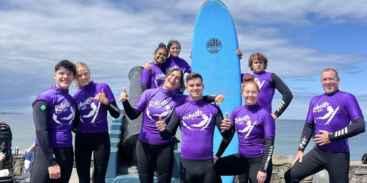 9 people wearing purple all in one swim tops. Standing and posing around a blue surf board. Stone beach and blue ocean in the background with blue sky and white clouds. 