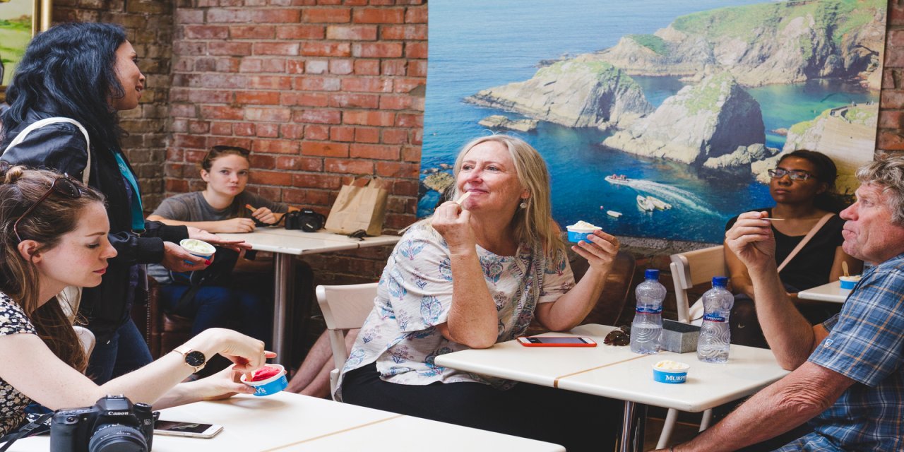 Red brick wall in the background with a large poster of the blue sea and cliffs handing on the wall. Four white tables can be seen with people sitting at them tasting food. 